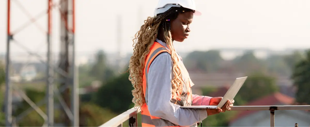 Field engineer wearing a hard hat and high-visibility safety vest standing on an elevated platform near telecommunications towers, using a laptop to review data outdoors.