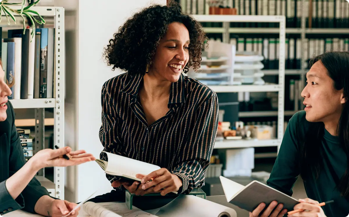 Three colleagues sitting together in an office, holding notebooks and engaged in a friendly discussion.