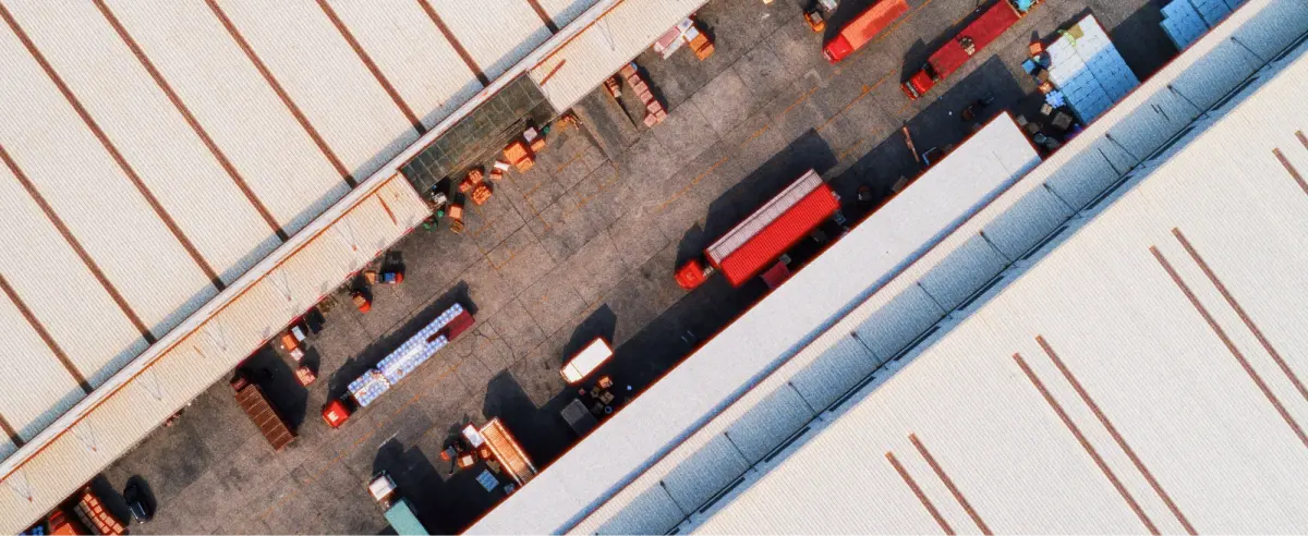 Aerial view of a warehouse with a corrugated roof, surrounded by red trucks and shipping containers. The scene is orderly and industrial.