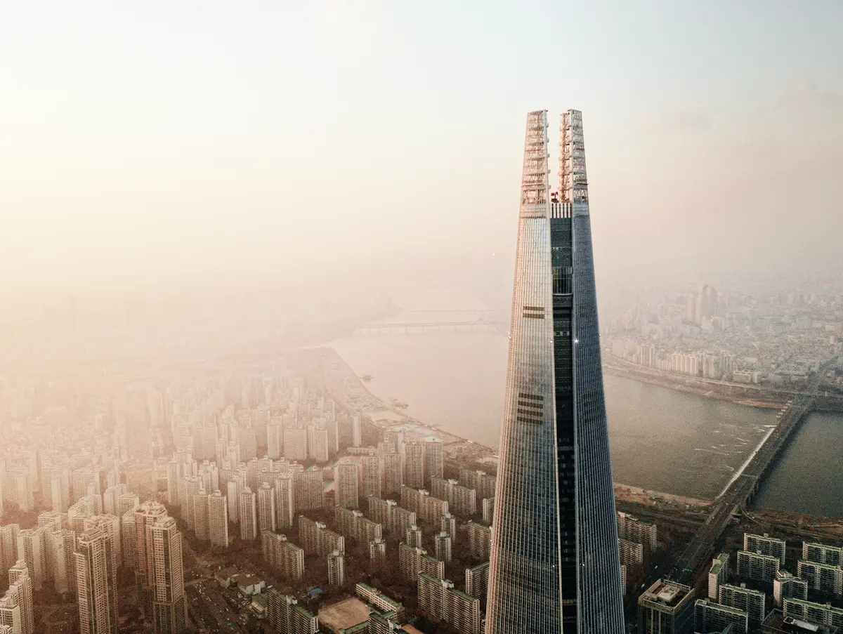 Aerial view of a tall modern skyscraper rising above a dense cityscape with a river and hazy skyline in the background.