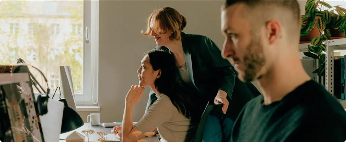 Three people working in an office, two collaborating at a computer while another focuses on his own work in the foreground.